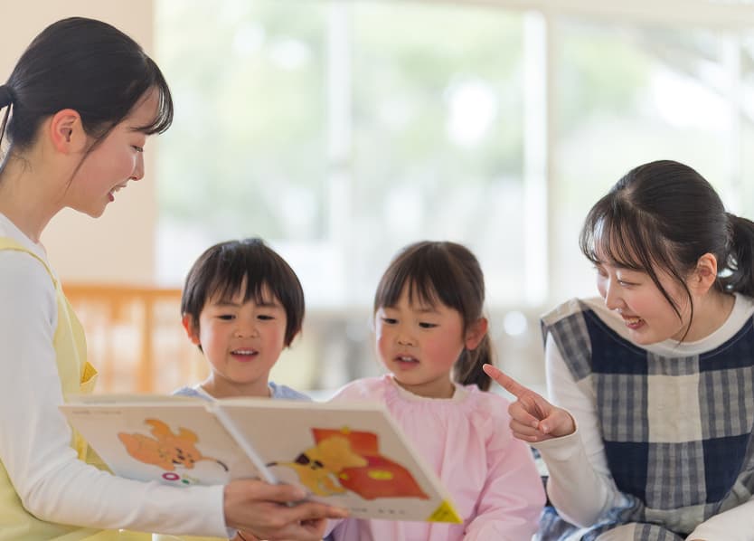 園内で子どもたちに先生が絵本の読み聞かせをしている画像です。